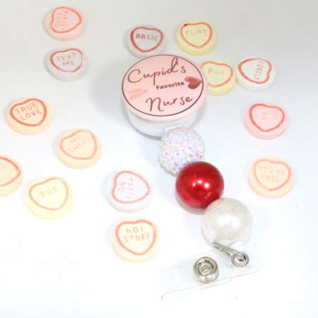 Cupid's Favorite Nurse badge reel with top circle in pink and red and bottom three beads. First bead is bumpty glitter white, second bead is shiny red and third bead is matte white. Surrounded by candy hearts.