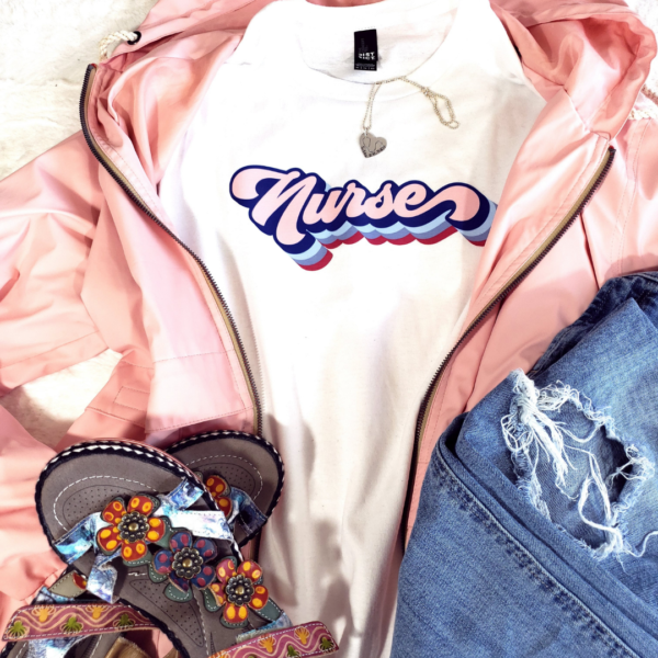 Pink jacket open with white tshirt underneath that says Nurse in a retro font with jeans and sandals and a heart necklace